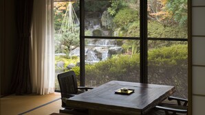 Ein Zimmer mit Tatami-Boden, einem Holztisch mit Kissen, einem Stuhl und Blick auf einen Wasserfall durch große Fenster.