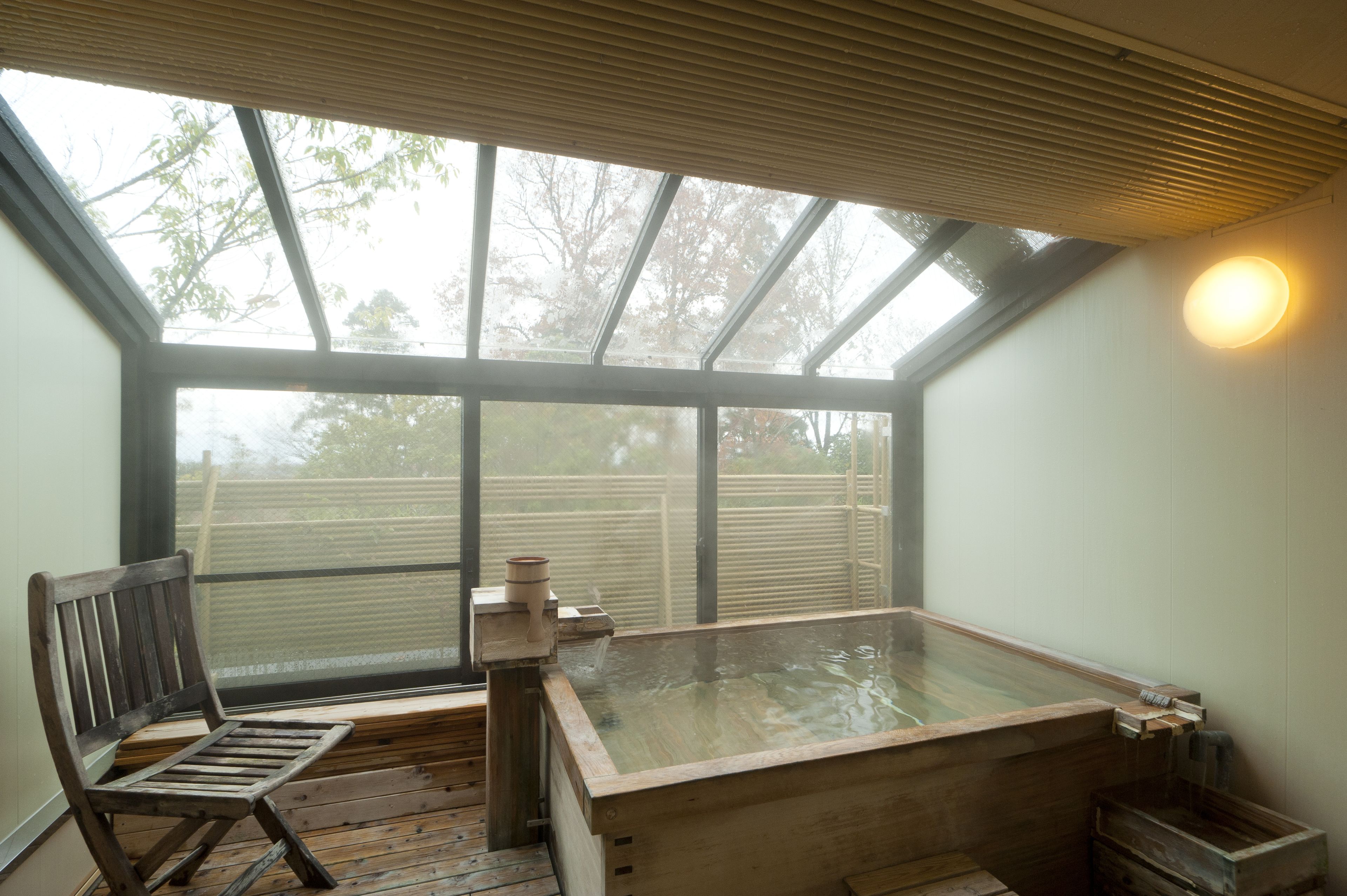 A wooden bathtub in a room with a large window, a wooden chair, and a wooden bench.