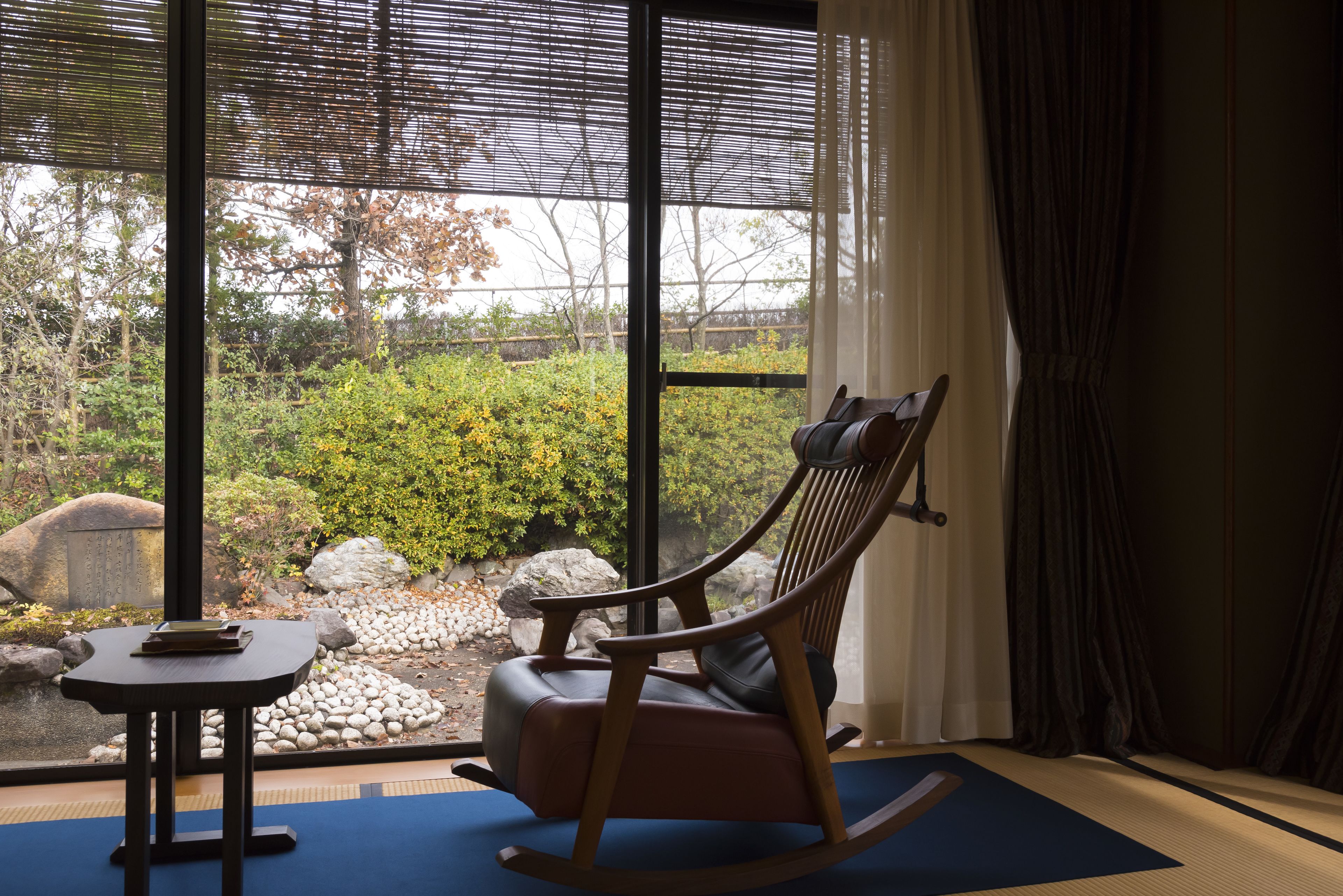 Ein Zimmer mit Schaukelstuhl, einem kleinen Tisch und Blick auf einen Garten durch große Fenster.