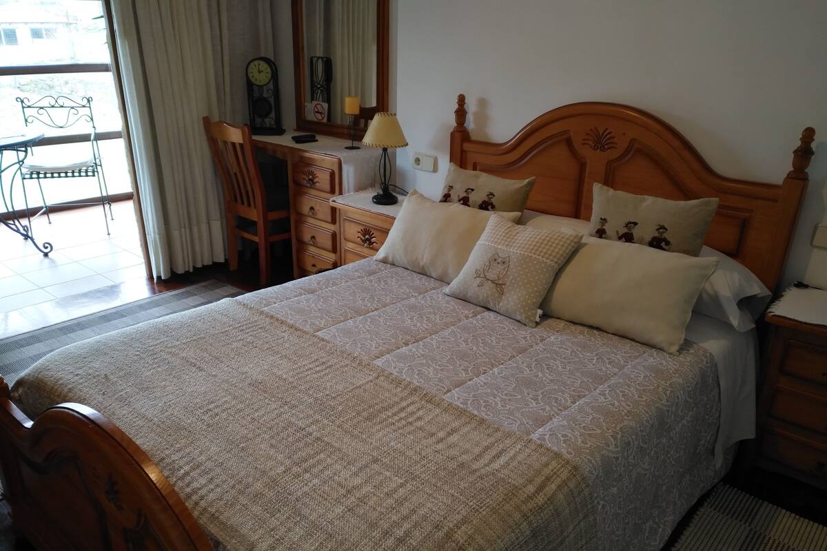Quarto com cama grande, cabeceira de madeira, escrivaninha com cadeira, luminária e janela com cortinas.