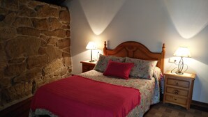 Un dormitorio con pared de piedra, una cama de madera con ropa de cama floral, una manta roja, dos lámparas de noche y una mesita de noche de madera con cajones.