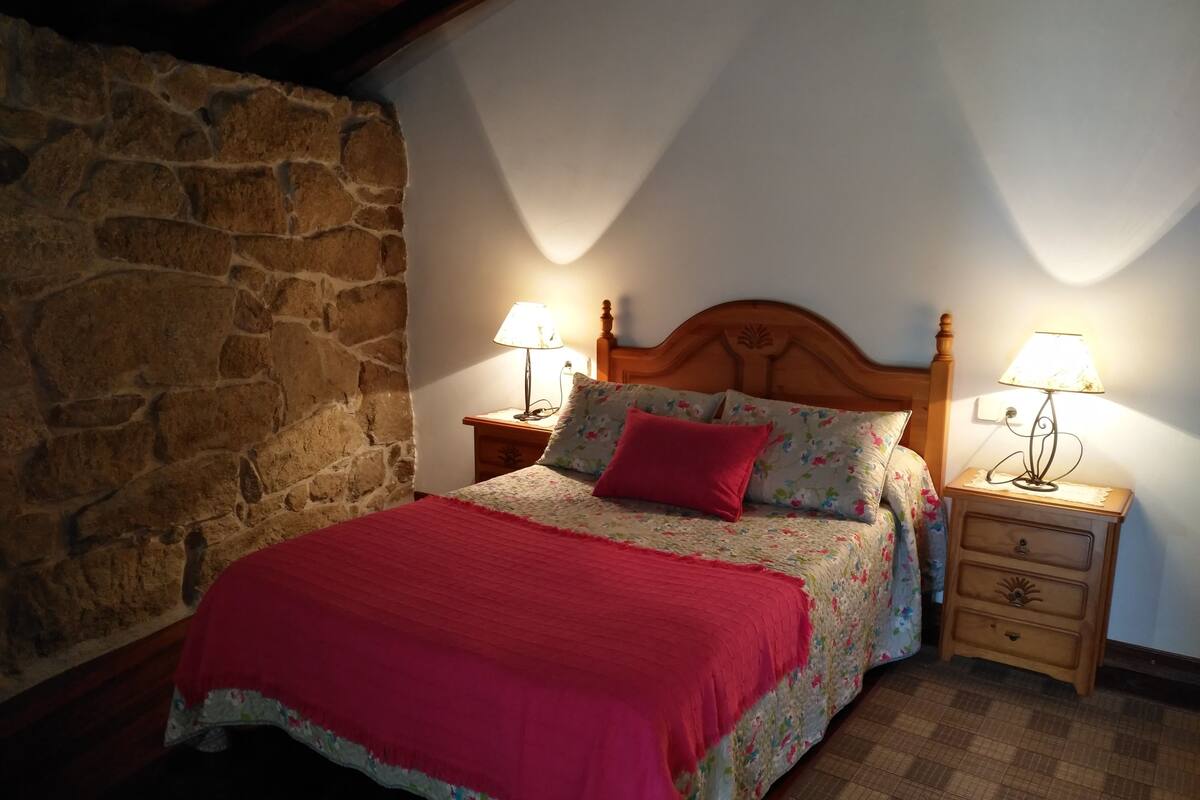Quarto com parede de pedra, cama de madeira com roupa de cama floral, manta vermelha, duas luminárias de cabeceira e mesa de cabeceira de madeira com gavetas.
