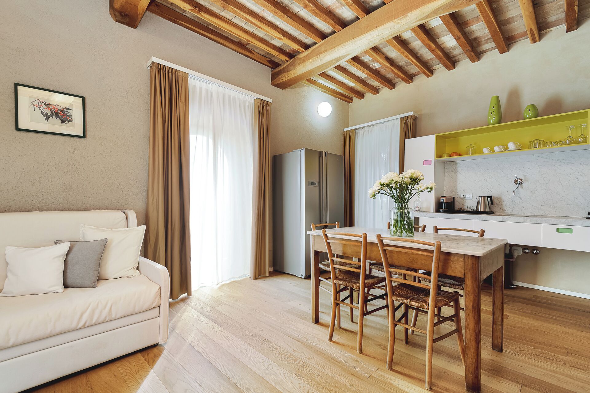 A cozy living room with a sofa, wooden dining table, and chairs. A kitchen area with a refrigerator and a shelf with glassware is visible.