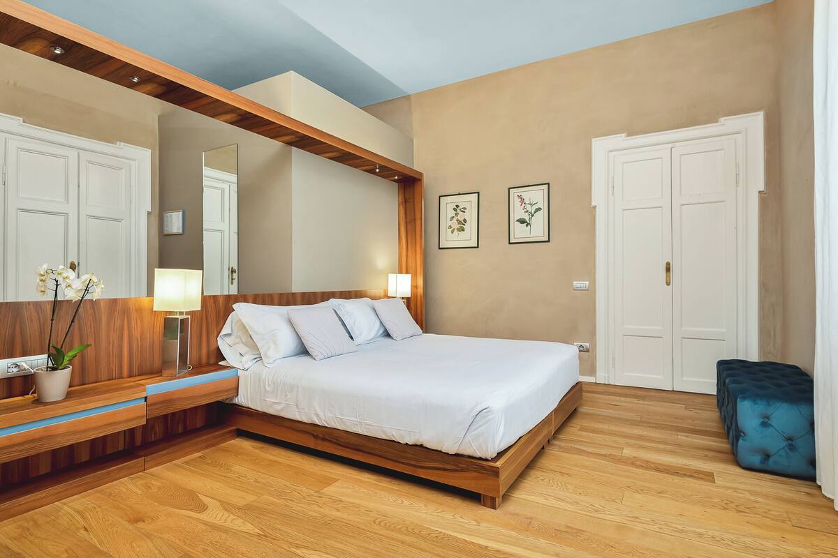 A bedroom with a wooden bed frame, white bedding, a nightstand with a lamp, a potted plant, and framed botanical prints on the wall.