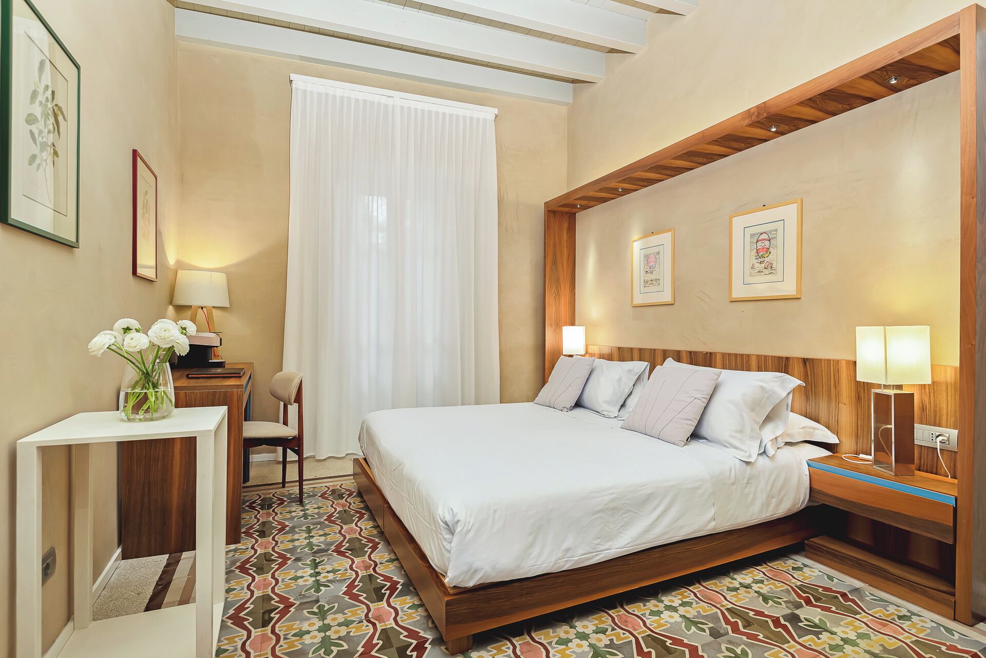 A hotel room with a large bed, a wooden headboard, a small desk with a lamp, a chair, and a window with white curtains.