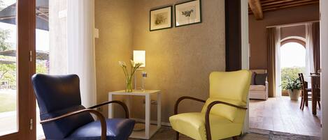 Una habitación con suelo de piedra, dos sillones, una mesita con lámpara y cuadros enmarcados en la pared.