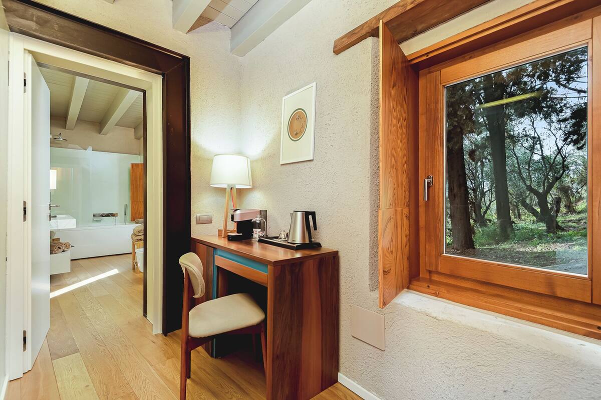 A wooden desk with a chair, a lamp, and a coffee maker in a room with a large window overlooking a forest.
