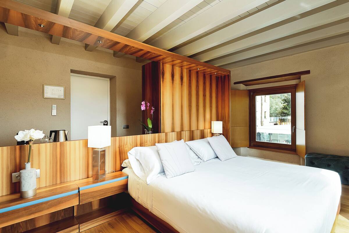 A hotel room with a large bed, wooden headboard, bedside tables, a mirror, a window with a view, and a small desk with a lamp.