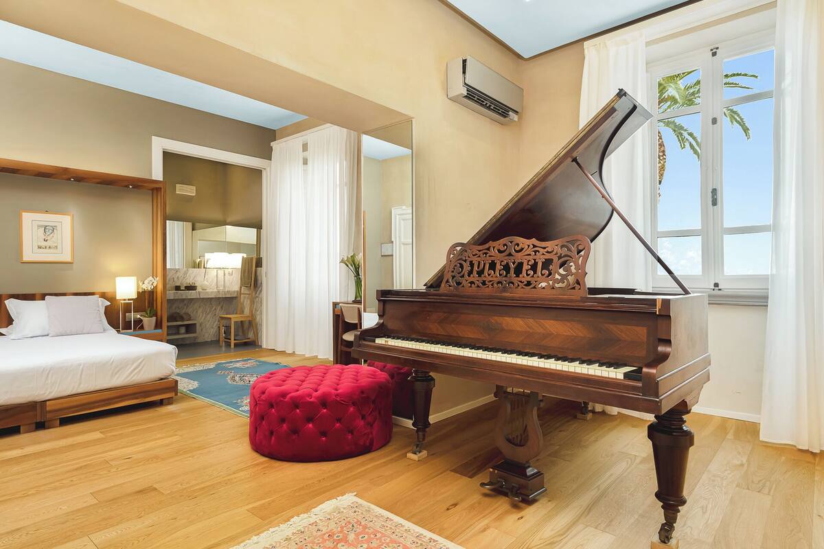 A hotel room with a grand piano, a bed, a red ottoman, a bathroom with a mirror, and a view of palm trees.