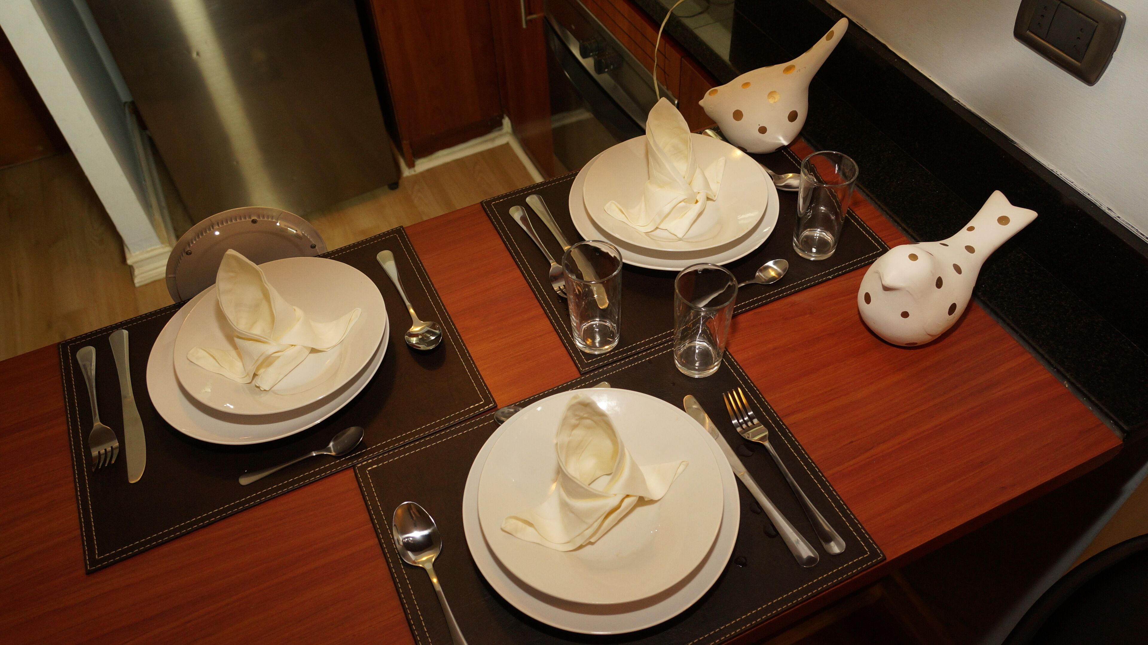 Una mesa de comedor dispuesta con platos, cubiertos y vasos, con una jarra de cerámica decorativa a la derecha.