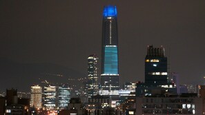 Une vue nocturne d’une ville, avec des gratte-ciel illuminés, dont un bâtiment particulièrement imposant dont le sommet est éclairé en bleu.