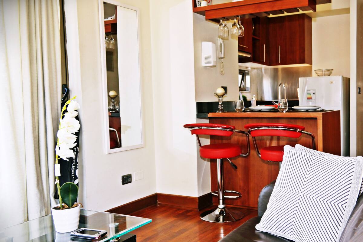 A compact living space with a kitchenette, a glass table with a smartphone, a potted plant, and a sofa with a patterned pillow.