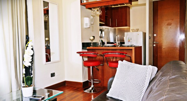Un espace de vie compact comprenant un coin cuisine, une table en verre avec un smartphone, une plante en pot et un canapé agrémenté d’un coussin à motifs.