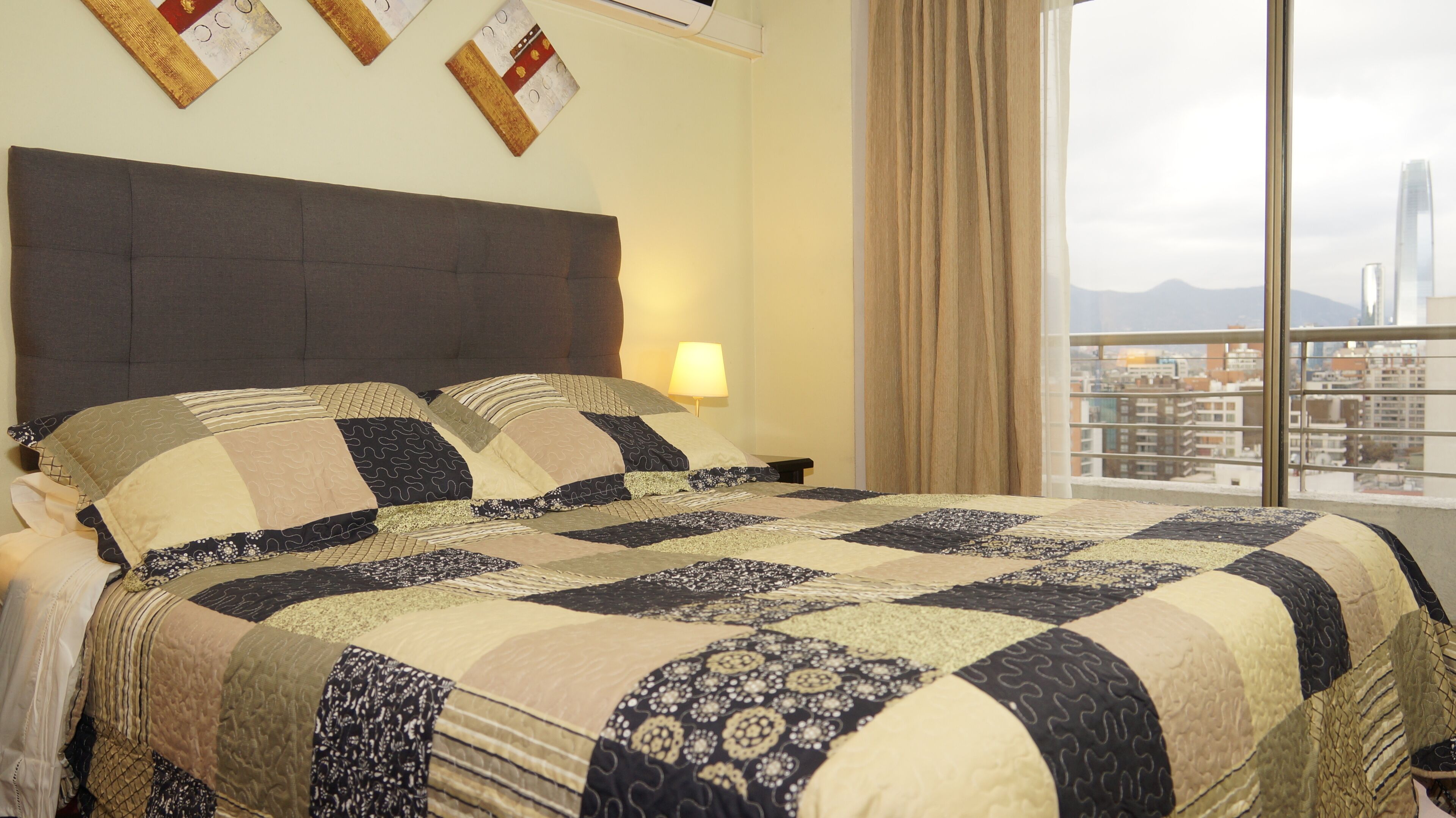 Habitación de hotel con una cama grande, cabecera, mesita de noche, lámpara y vista a un paisaje urbano a través de una ventana.