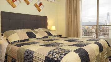 Habitación de hotel con una cama grande, cabecera, mesita de noche, lámpara y vista a un paisaje urbano a través de una ventana.