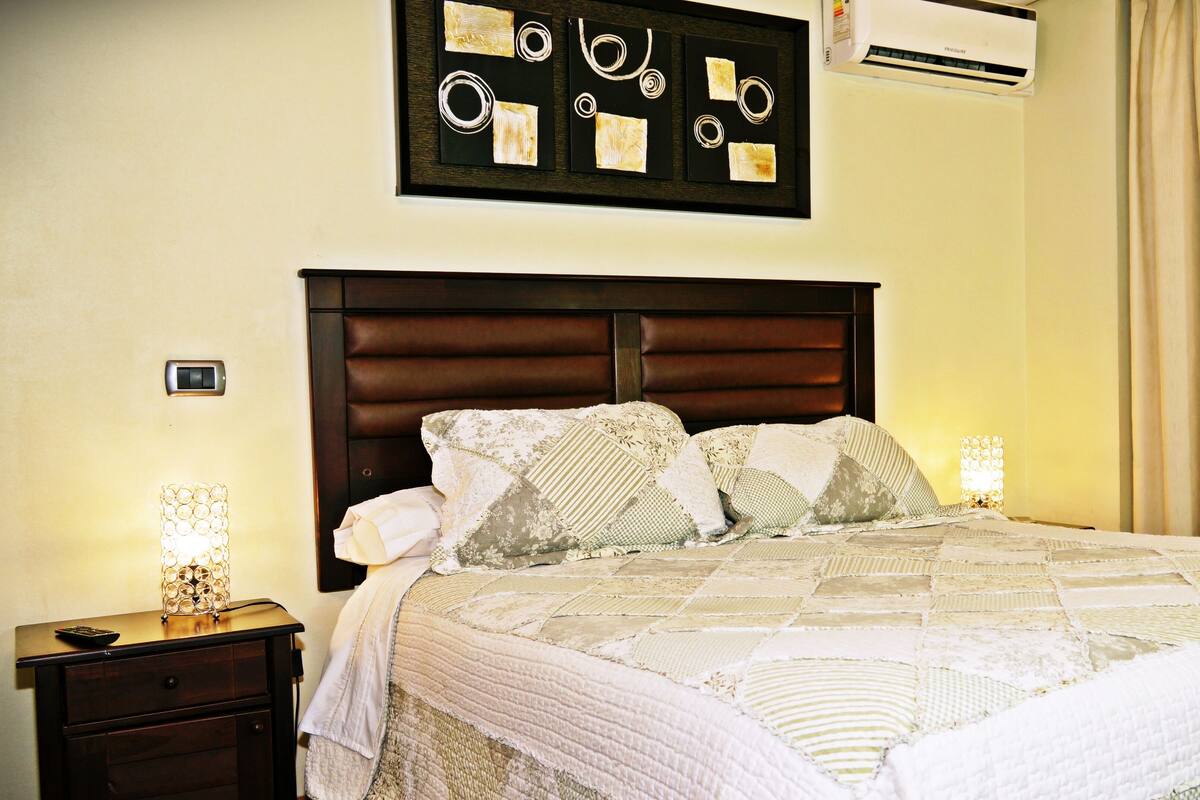 A neatly made bed with a quilt, a wooden headboard, a nightstand with a lamp, and a wall-mounted air conditioner.