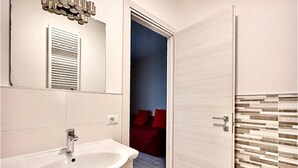 A modern bathroom with a white sink, a mirror, and a door leading to a bedroom with a red bedspread. - B&B Santa Maria Novella (Florence)