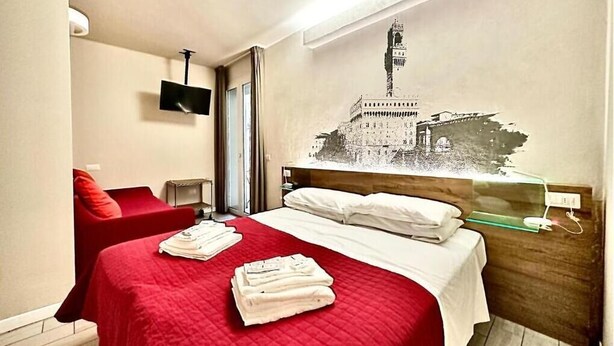 A hotel room with a bed, a red chair, a wall-mounted TV, and a wall art of a building. - B&B Santa Maria Novella (Florence)