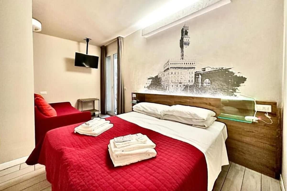 A hotel room with a bed, a red chair, a wall-mounted TV, and a wall art of a building.