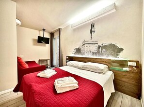 Une chambre d’hôtel avec un lit, un fauteuil rouge, un téléviseur fixé au mur et une œuvre d’art représentant un bâtiment.