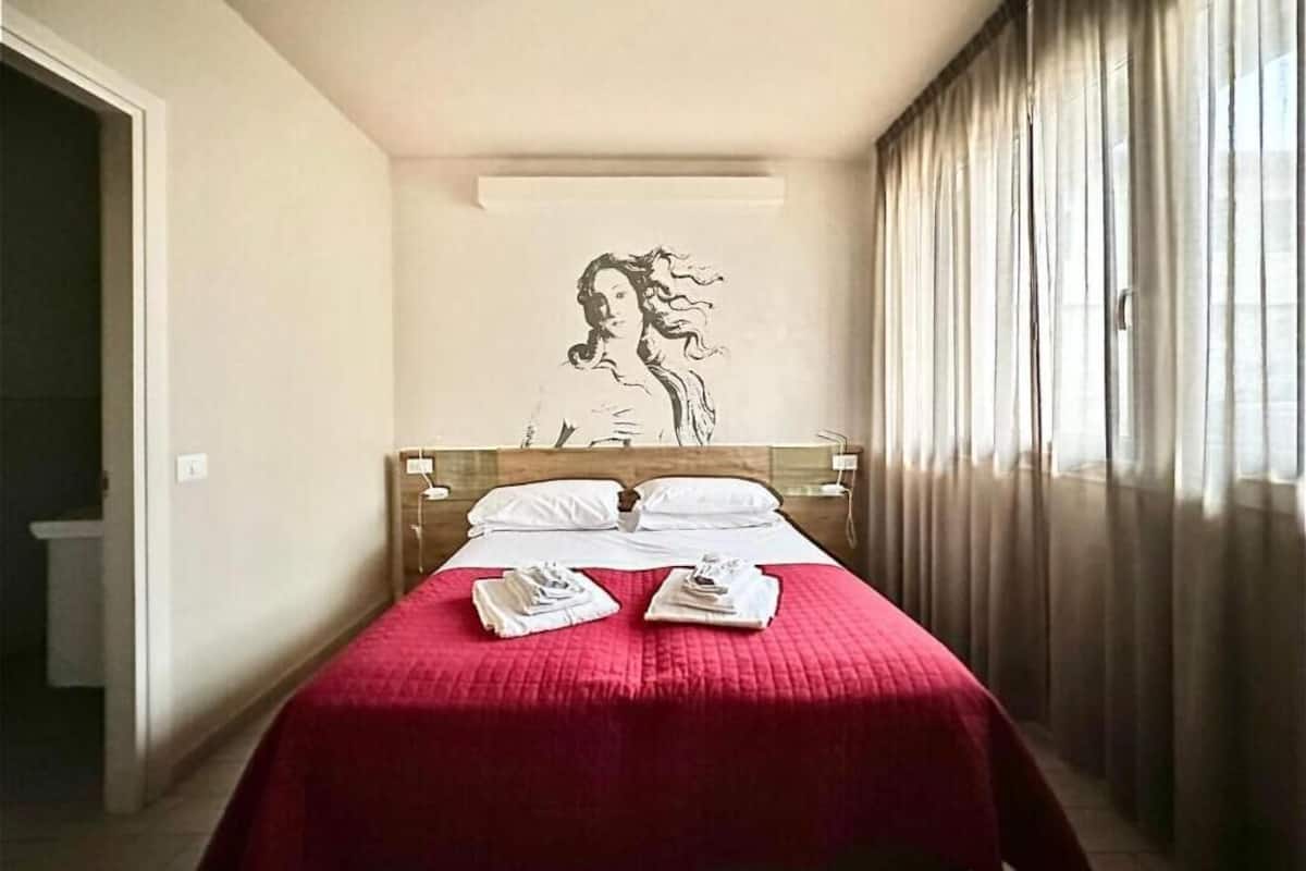 A hotel room with a bed, a headboard, a wall-mounted artwork, and a window with curtains.