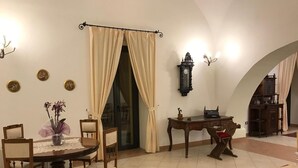 Un comedor con una mesa redonda, sillas y un florero. Hay un reloj en la pared y una ventana con cortinas.