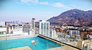 A rooftop pool with a glass barrier, overlooking a cityscape and a hill. - Apartamentos Nueva Providencia 1372 (Santiago)