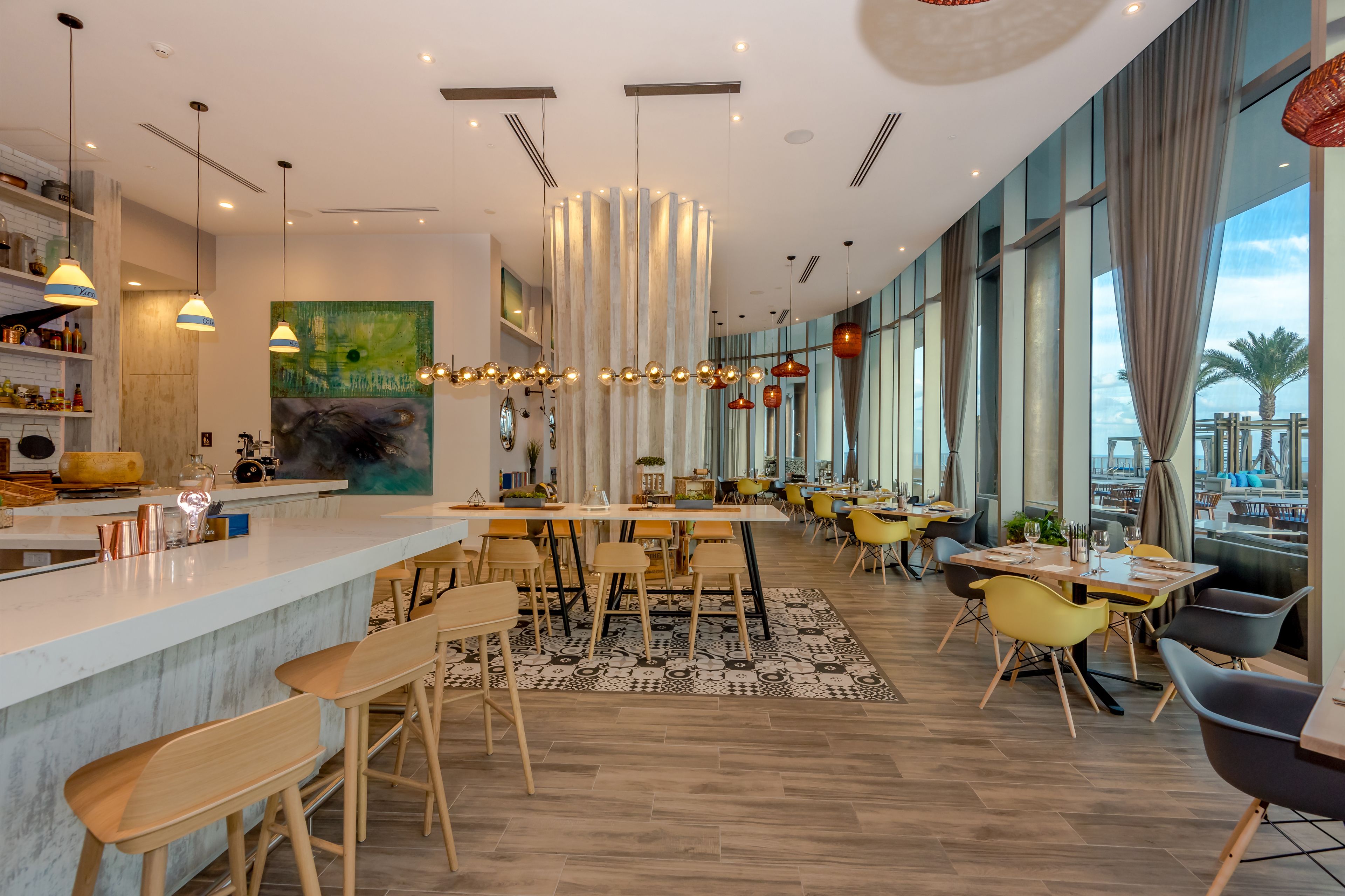 A modern restaurant with a bar area, wooden flooring, and large windows offering a view of the outdoors.