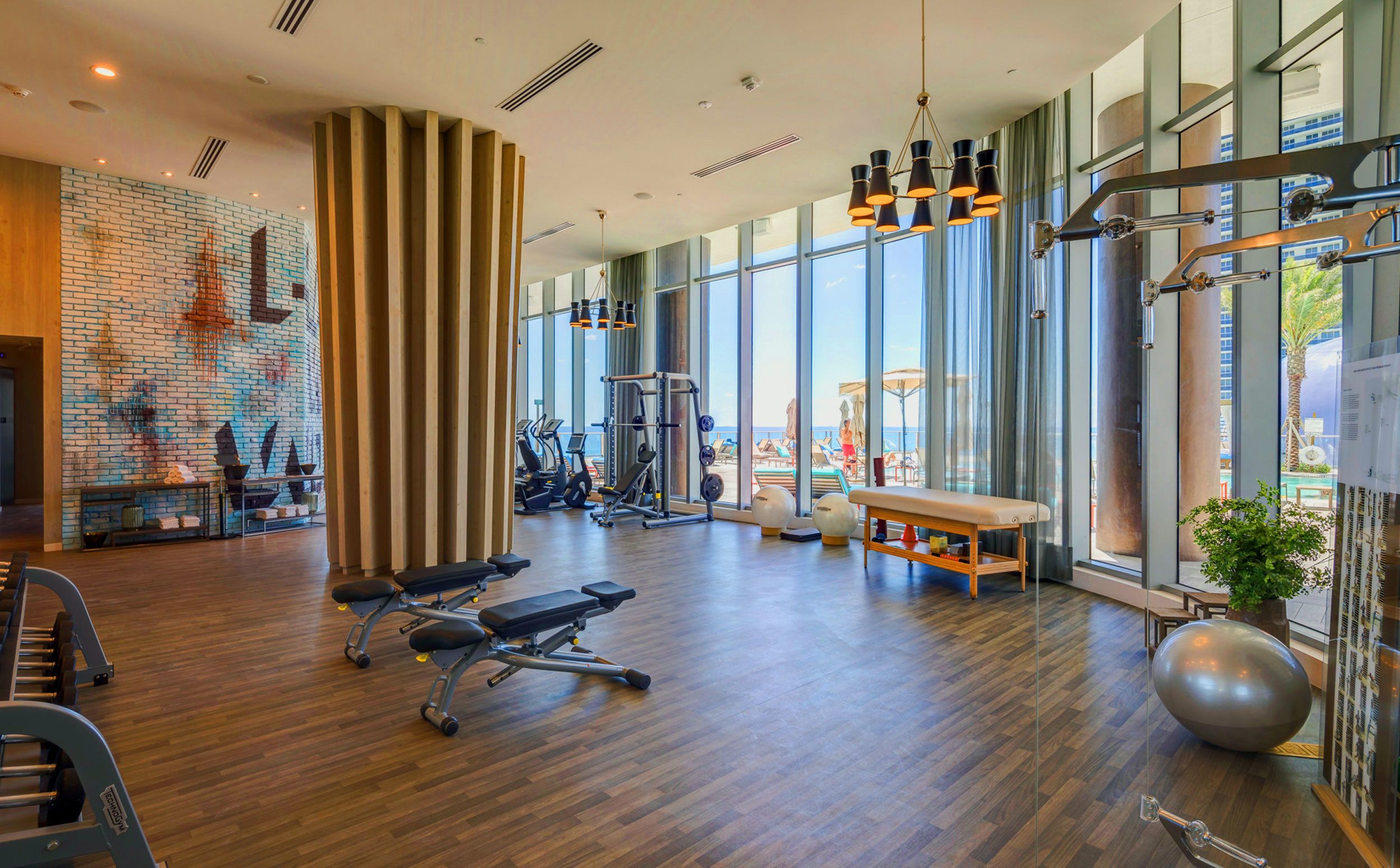 Une salle de sport spacieuse dotée d’un plancher en bois, d’équipements d’exercice et de grandes fenêtres offrant une vue sur la plage.