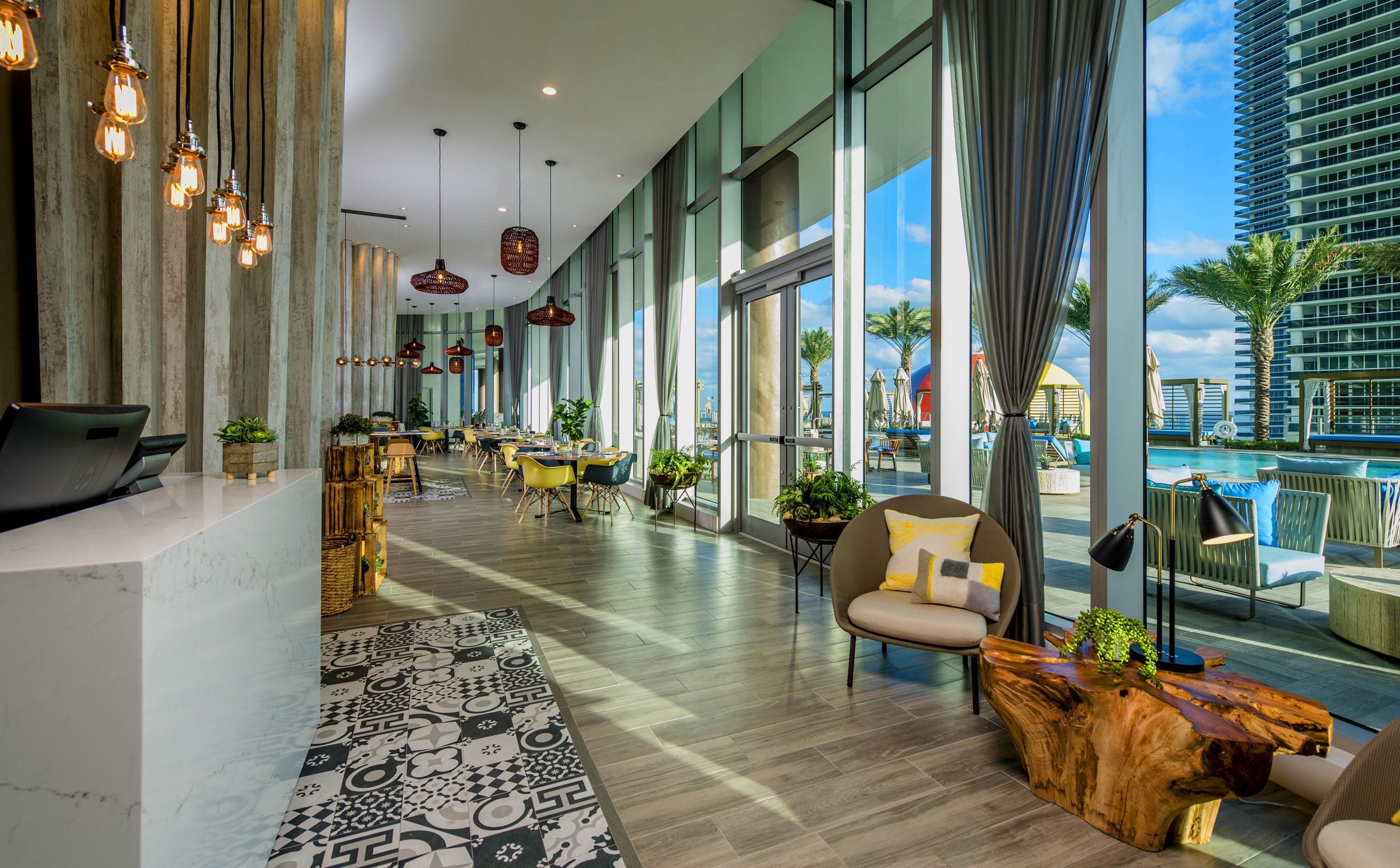 A modern hotel lobby with a pool view, featuring a reception desk, seating area, and large windows.