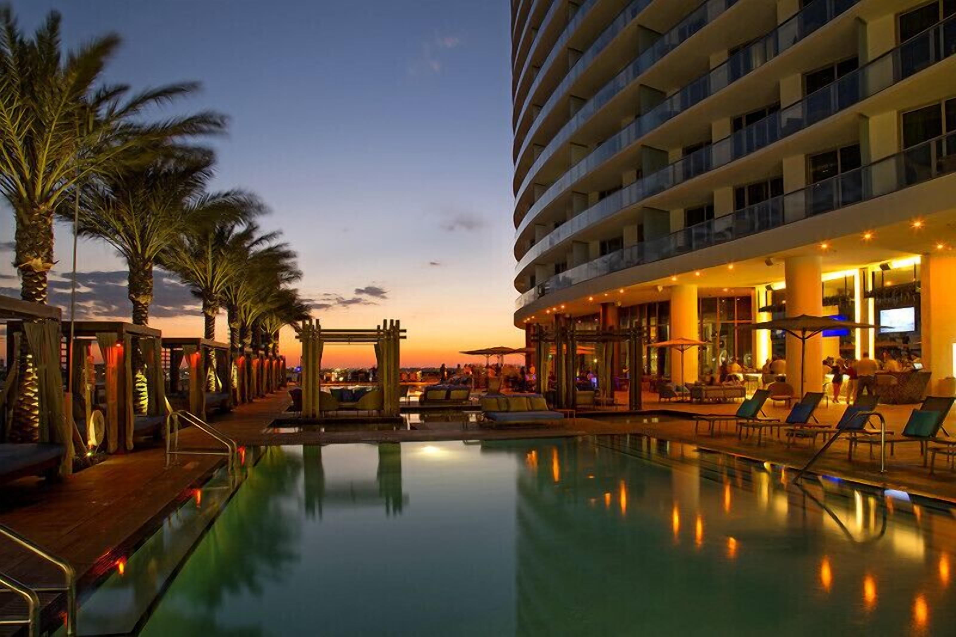 A hotel pool area with a sunset view, palm trees, and a multi-story building with balconies.
