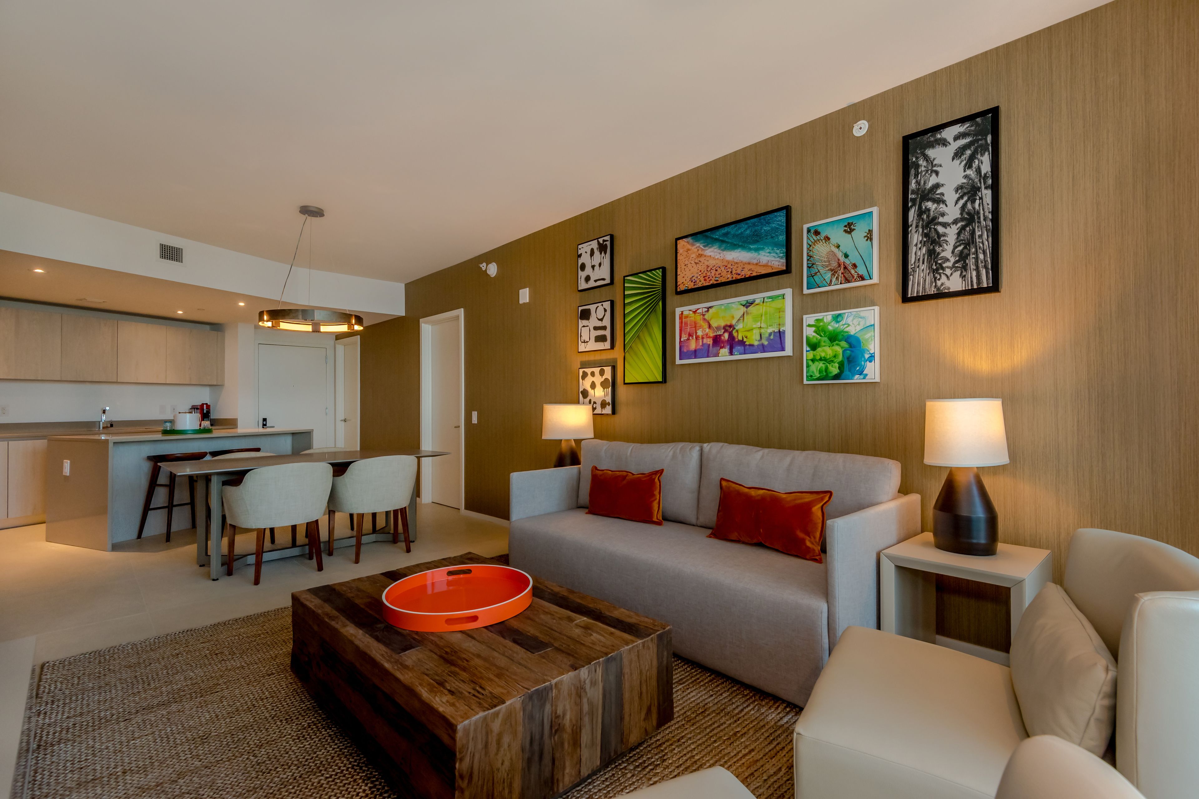 A modern living room with a sofa, coffee table, and a dining area with chairs. There are wall-mounted picture frames and a lamp on a side table.