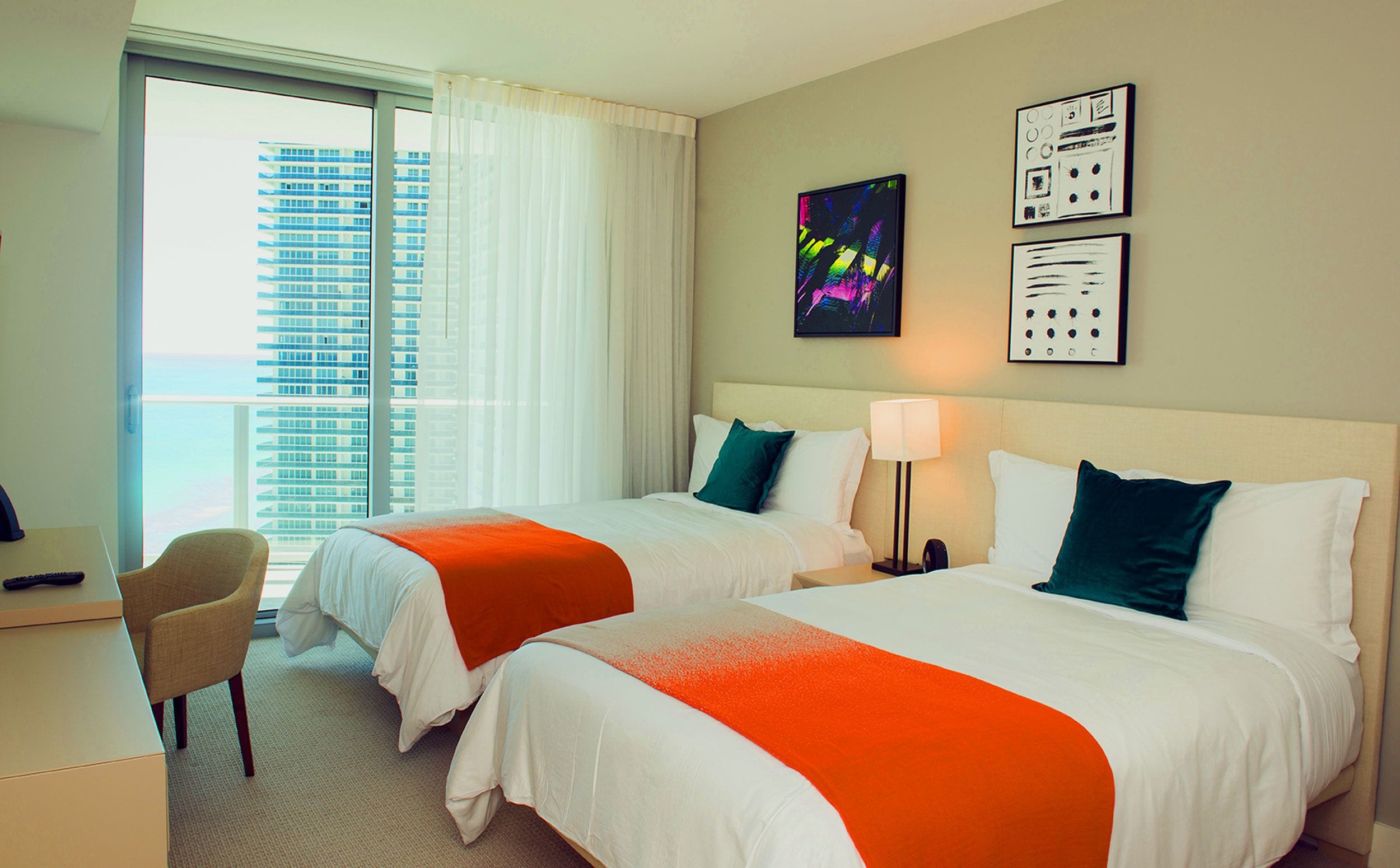 A hotel room with two beds, a desk, and a chair. There is a window with a view of a tall building.