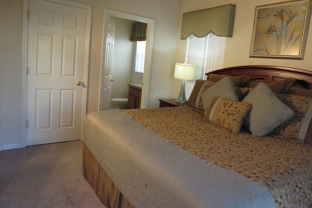 A bedroom with a bed, a nightstand, a lamp, a door, and a painting on the wall.