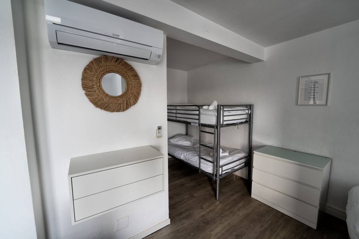 Une petite chambre avec des lits superposés, une commode blanche, un miroir et un appareil de climatisation.