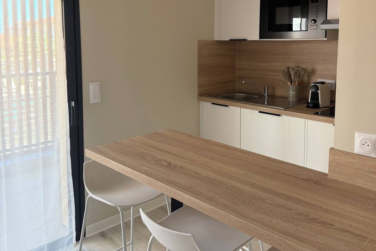 Une cuisine moderne avec un plan de travail en bois, des armoires blanches, un four à micro-ondes encastré et une cuisinière noire. On y trouve deux tabourets de bar blancs et une porte-fenêtre coulissante donnant sur un balcon.