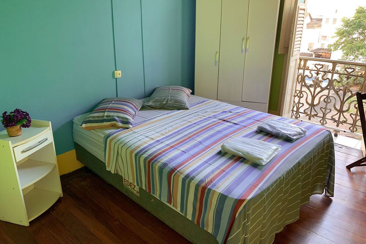 Un lit simple avec une couvre-lit à rayures et des oreillers, une petite table de chevet avec une plante, et une armoire.