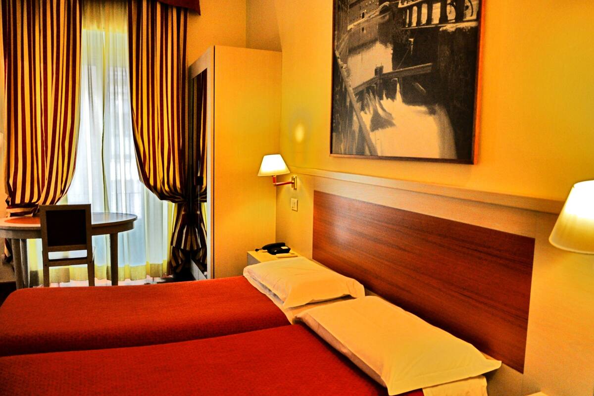 Ein Hotelzimmer mit zwei Betten, einem Schreibtisch, einem Stuhl, einer Lampe und einem Bild an der Wand.