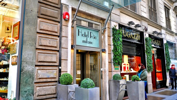 Entrada de un hotel con un cartel que dice "Hotel Rio Milano", una fachada con la señalización "UGAZZI" y una acera para peatones.