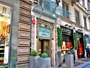 A hotel entrance with a sign reading "Hotel Rio Milano," a storefront with "UGAZZI" signage, and a pedestrian walkway. - Hotel Rio (Milan)