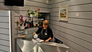 Un área de bar moderna con una barra de acero inoxidable, estantes con botellas y flores, y un cuadro en la pared.