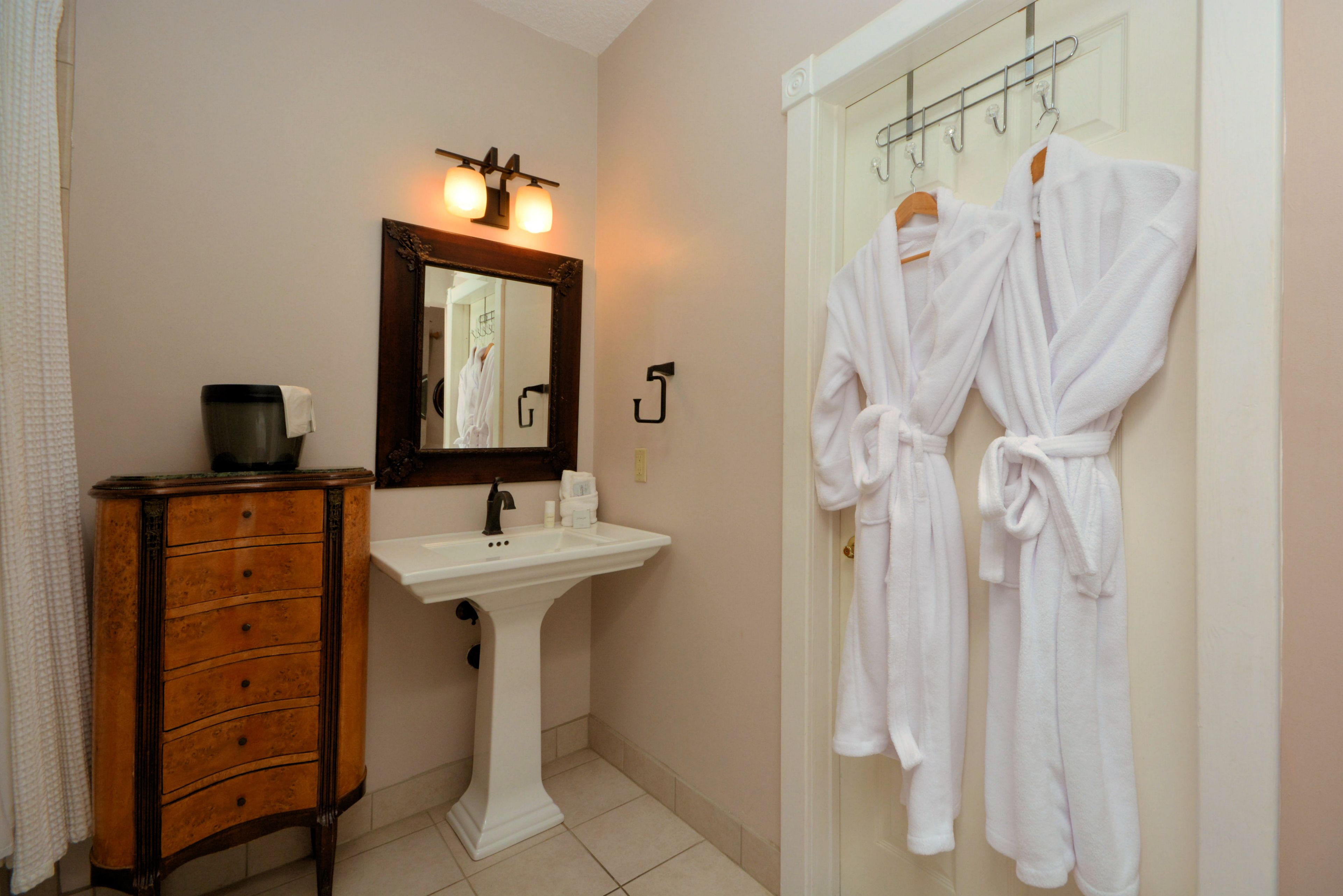 A bathroom with a wooden cabinet, a pedestal sink, and two white robes hanging on hooks.