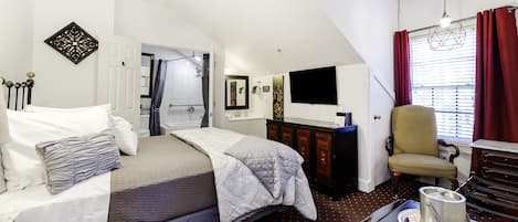 Ein Schlafzimmer mit einem Bett, einem Schreibtisch mit Wasserkocher, einem Sessel, einem Fernseher und einem Fenster mit Vorhängen.