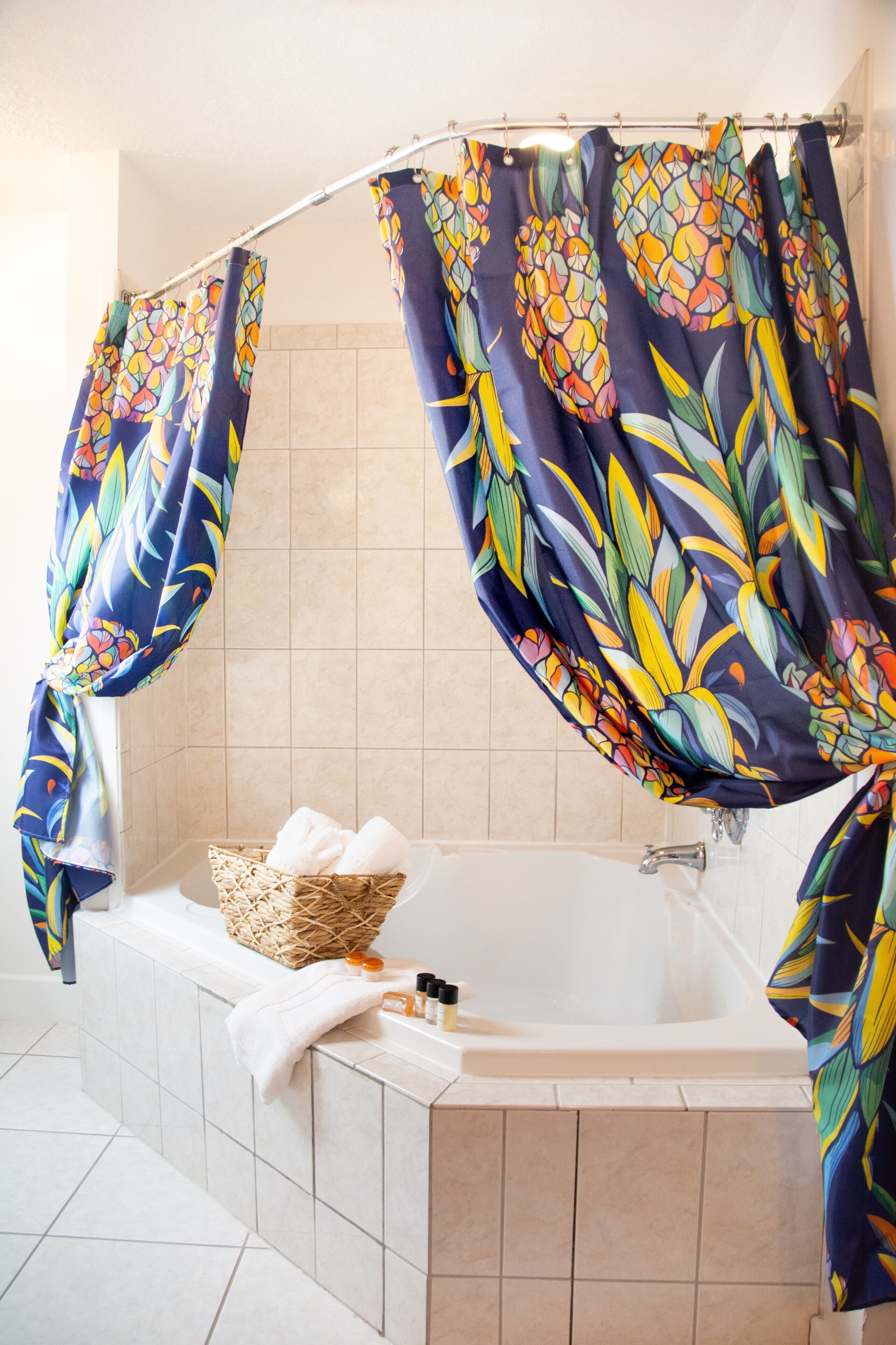 Un baño con una bañera, una cesta de mimbre con toallas y una cortina de ducha con estampado de hojas tropicales.