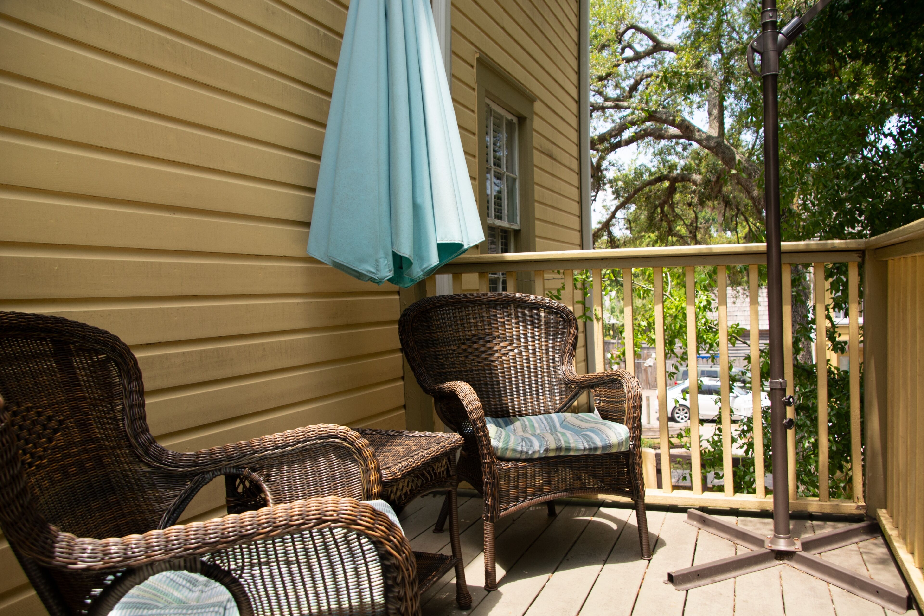 A porch with wicker chairs and a table, a blue umbrella, and a view of parked cars and trees.