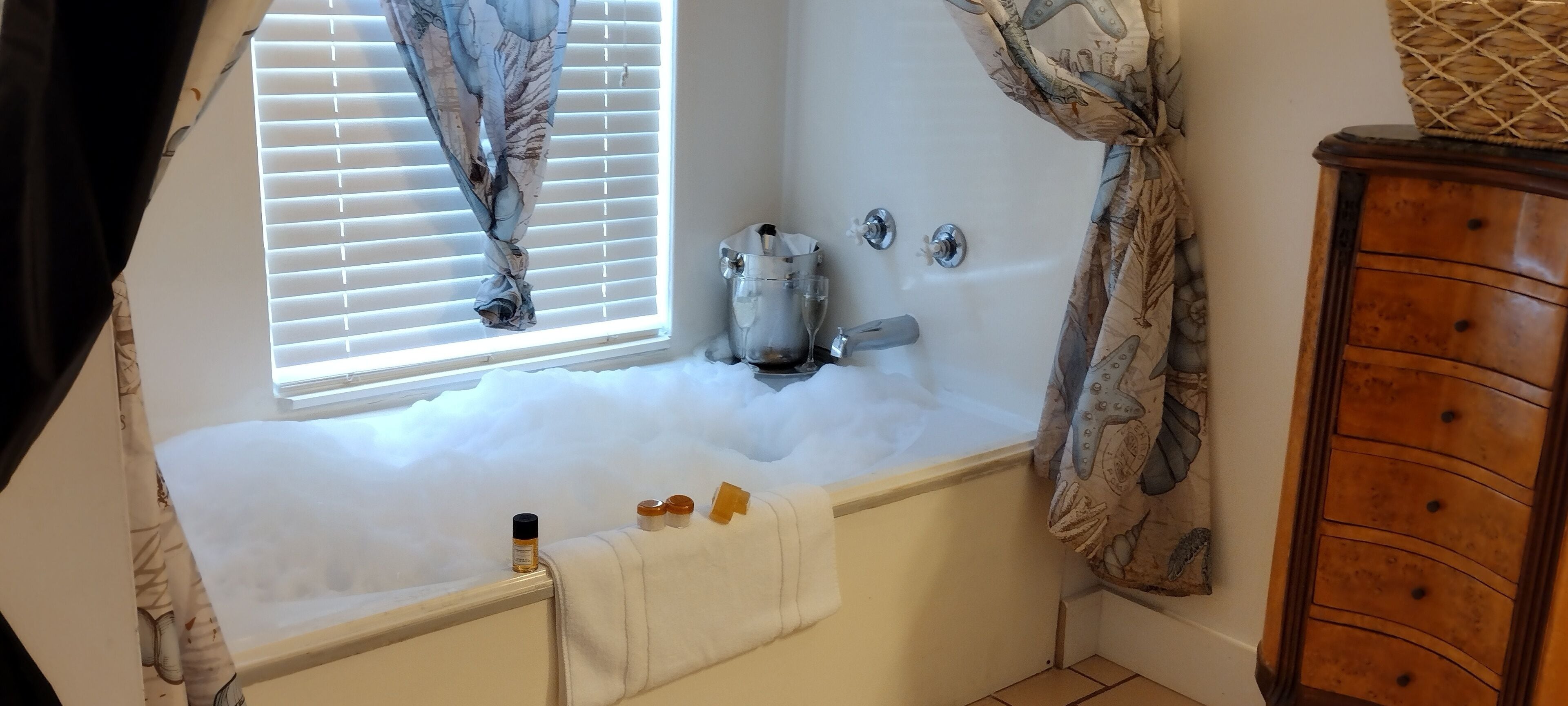 A bathroom with a bathtub, a window with curtains, a wooden cabinet, and toiletries on the edge of the tub.