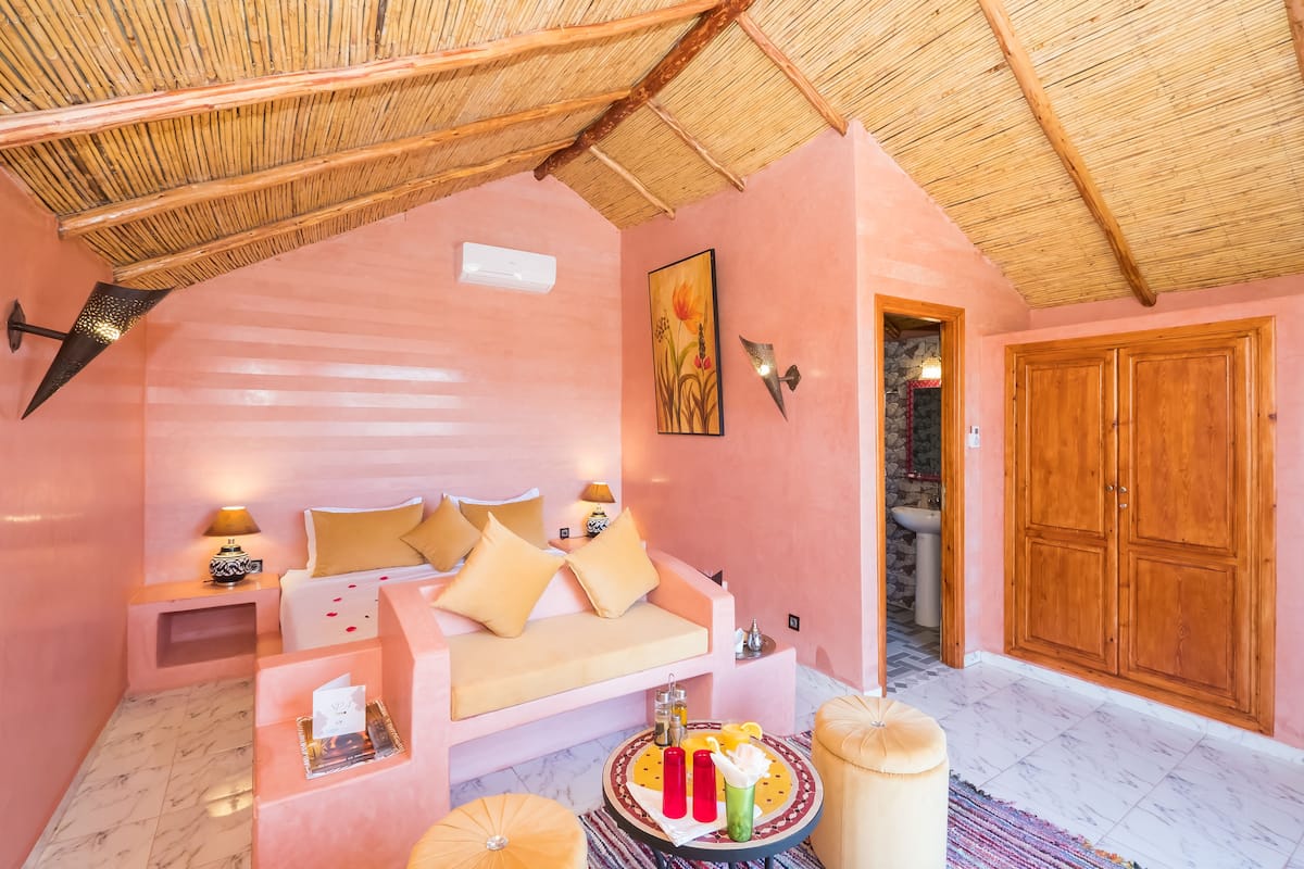 A pink-themed room with a thatched roof, a sofa with cushions, a bedside table with a lamp, a round table with drinks, and a wooden wardrobe.