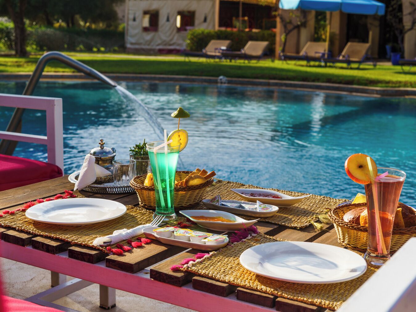 A poolside dining setup with a table, plates, glasses, and a water fountain.