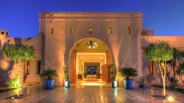 A hotel entrance with an arched doorway, potted plants, and a tiled pathway leading to the entrance. - Villa Marco Marrakech (Tameslouht)