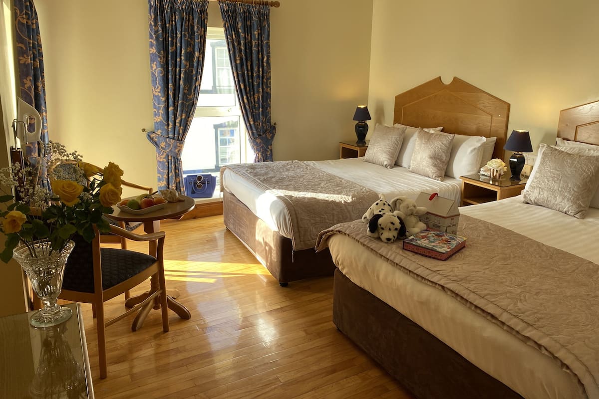 Habitación de hotel con dos camas, piso de madera, un ventanal grande con cortinas, una mesita de noche con lámpara y un jarrón con flores.